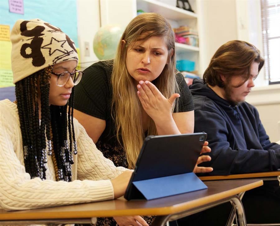  Two students in a classroom working on tablets. A teacher stands between them working a student.